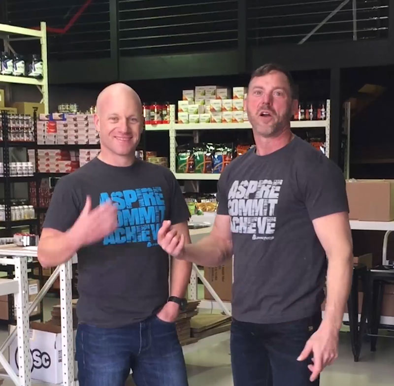 Tim and Mike standing in the SuppKings warehouse in front of racking stacked with supplements.